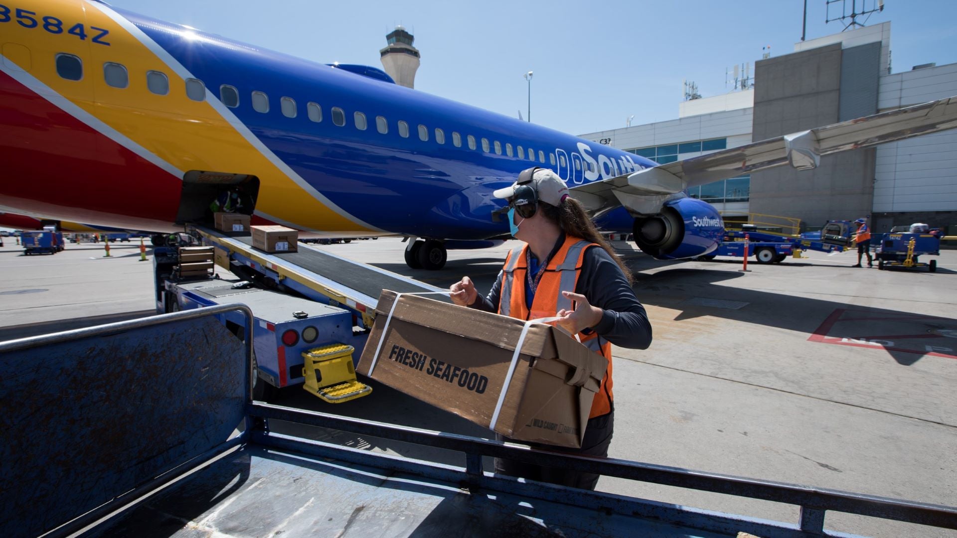 A female ground worker lifts a box from a cart onto a conveyor belt used to load a blue, yellow and red Southwest Airlines jet.