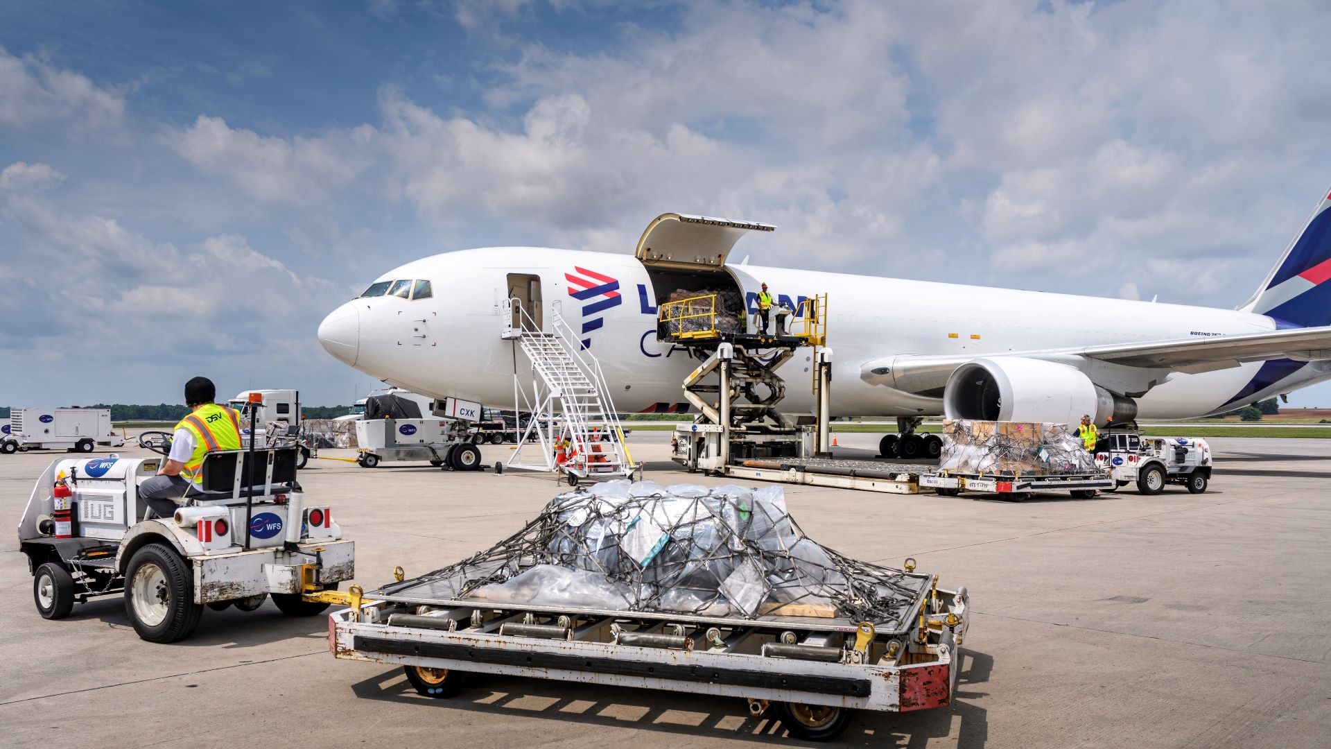 An airport tug pulls a cart with cargo toward a white jet with an open cargo door.