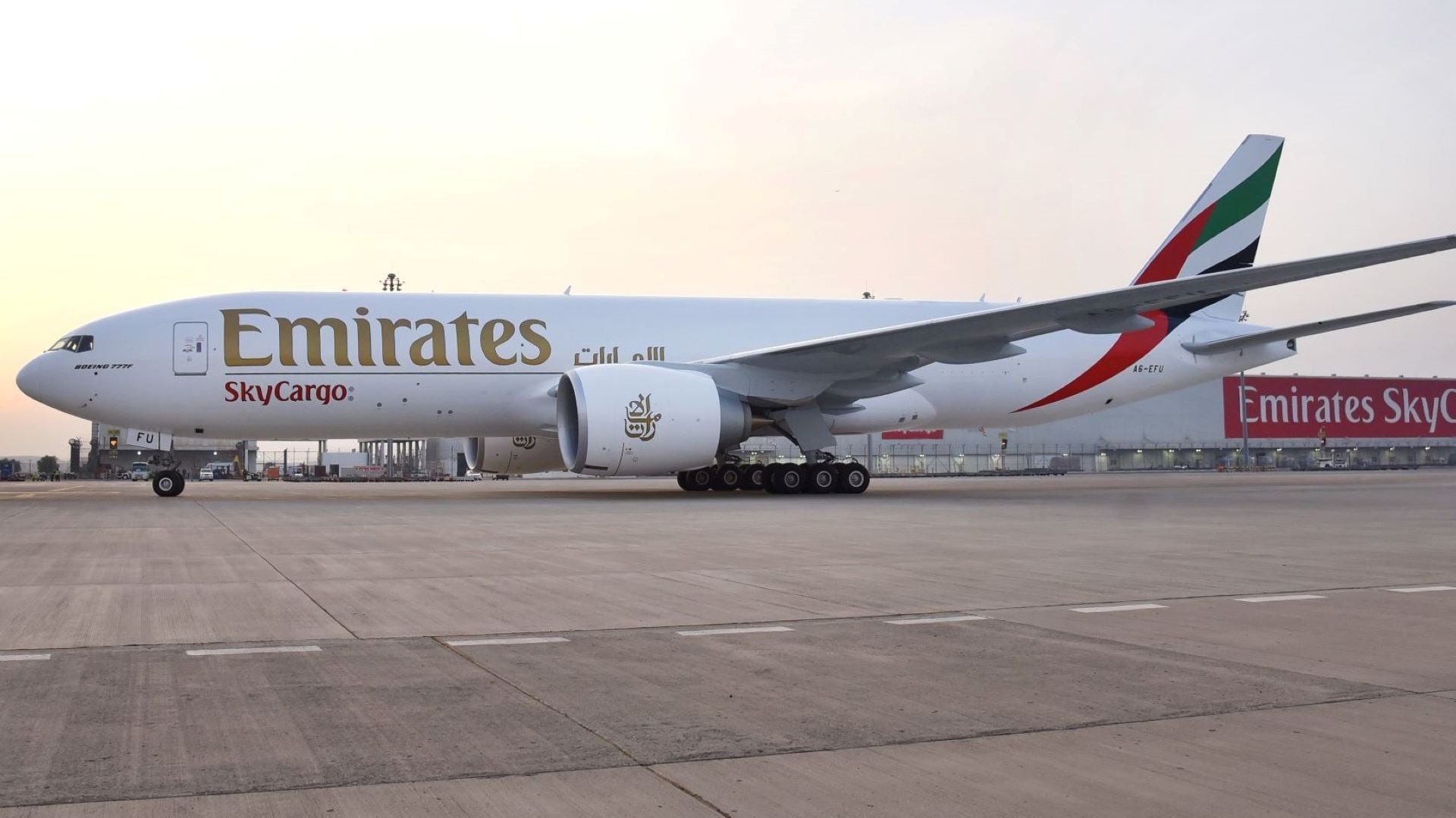 A white cargo jet with Emirates gold lettering and black/red coloring on tail passes camera on runway.