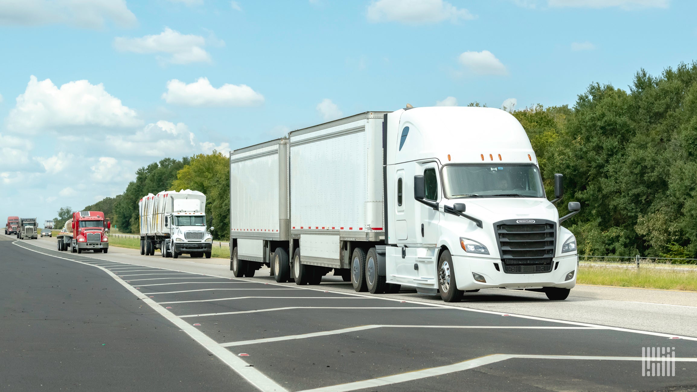 A white semi truck is followed by other trucks on a highway with green trees in the background.