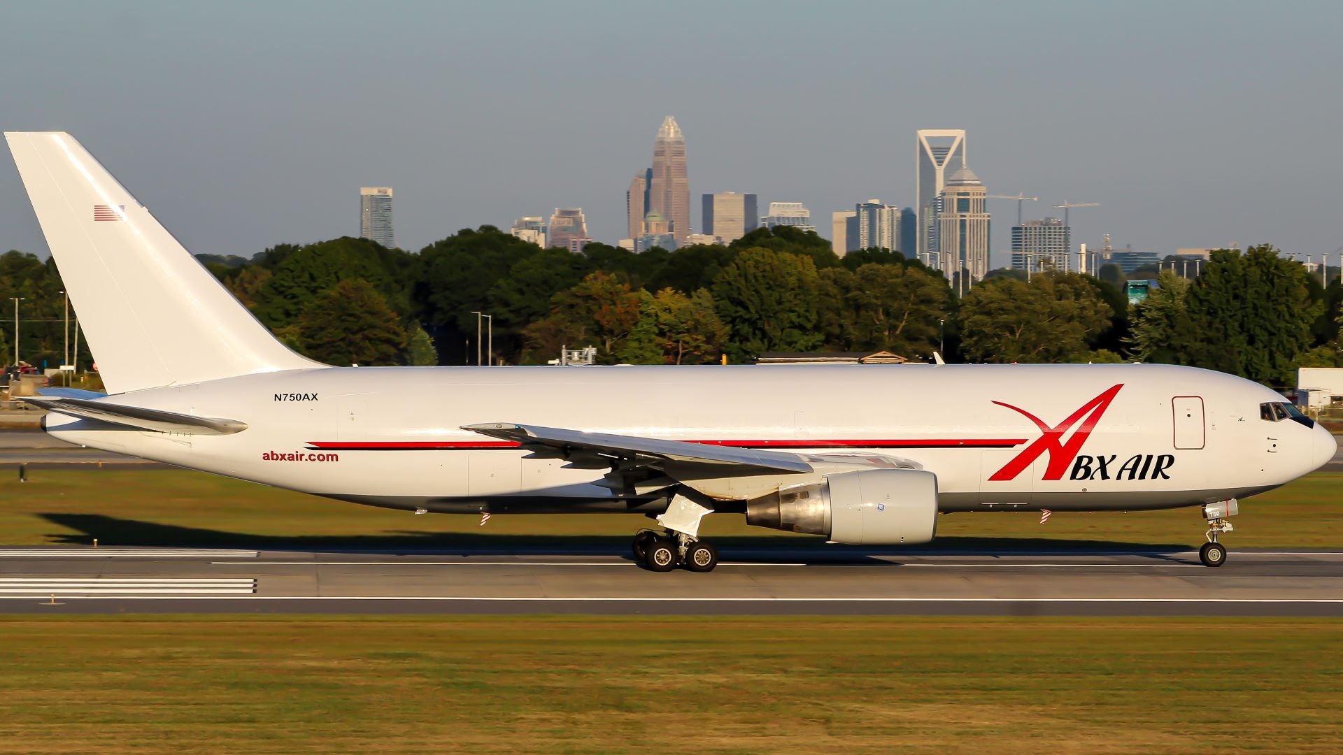 A large white jet with red accents and ABX Air logo on runway with trees and city skyline in background.