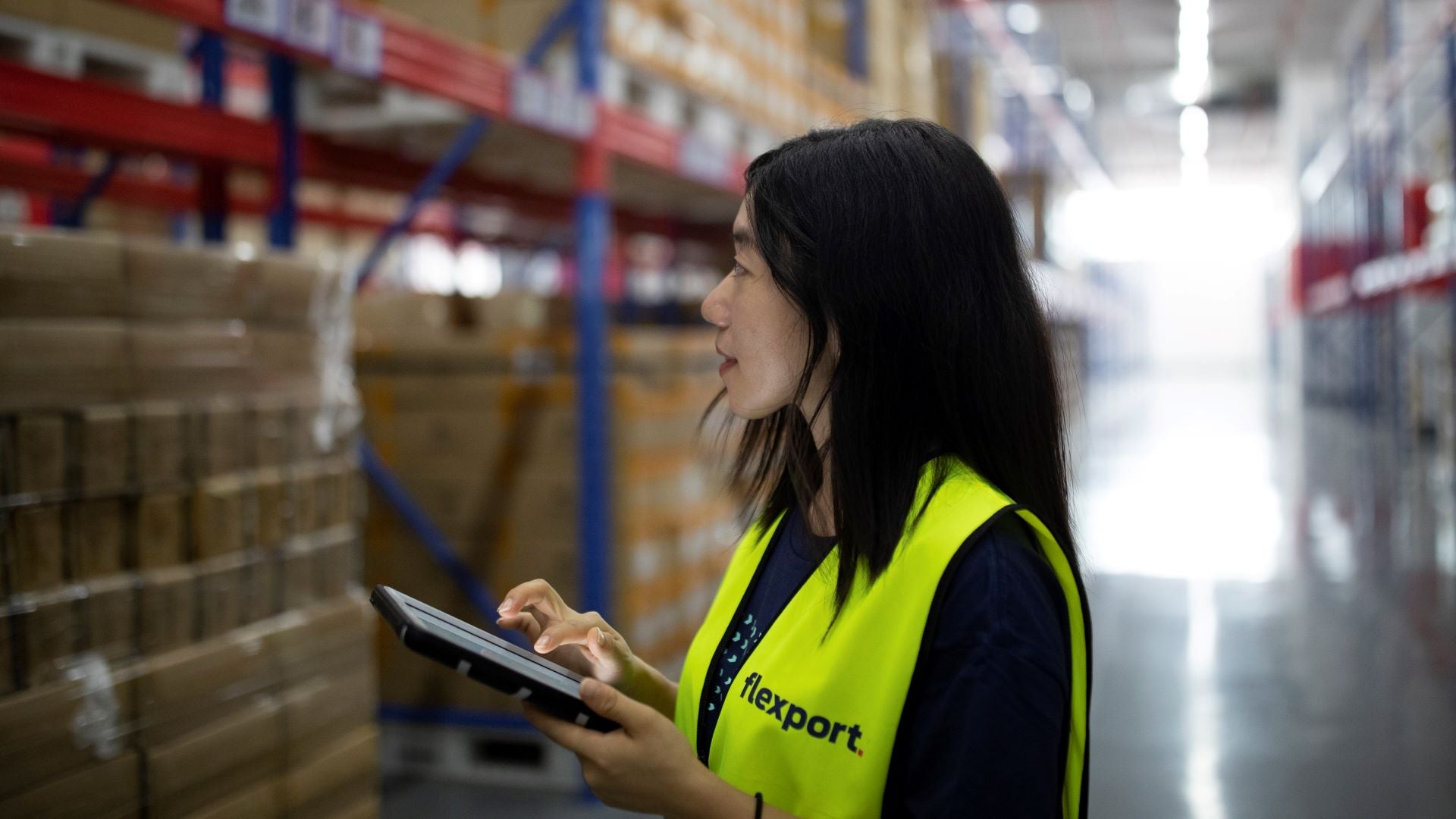 A woman with dark hair enters info into a digital pad while in warehouse. She is wearing a yellow safety vest.