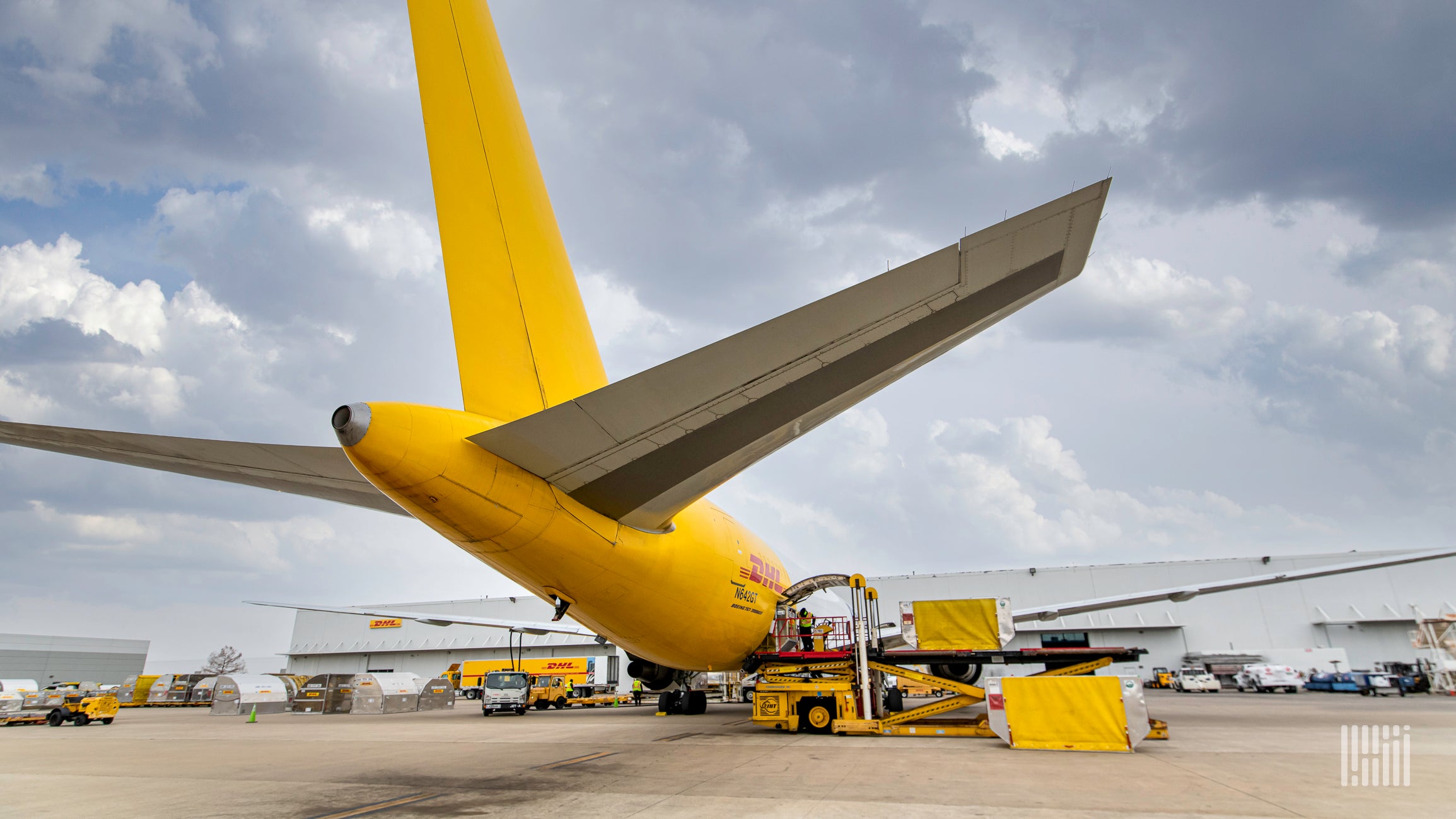 A mustard-yellow DHL plane loading cargo at an airport. View from behind the plane.