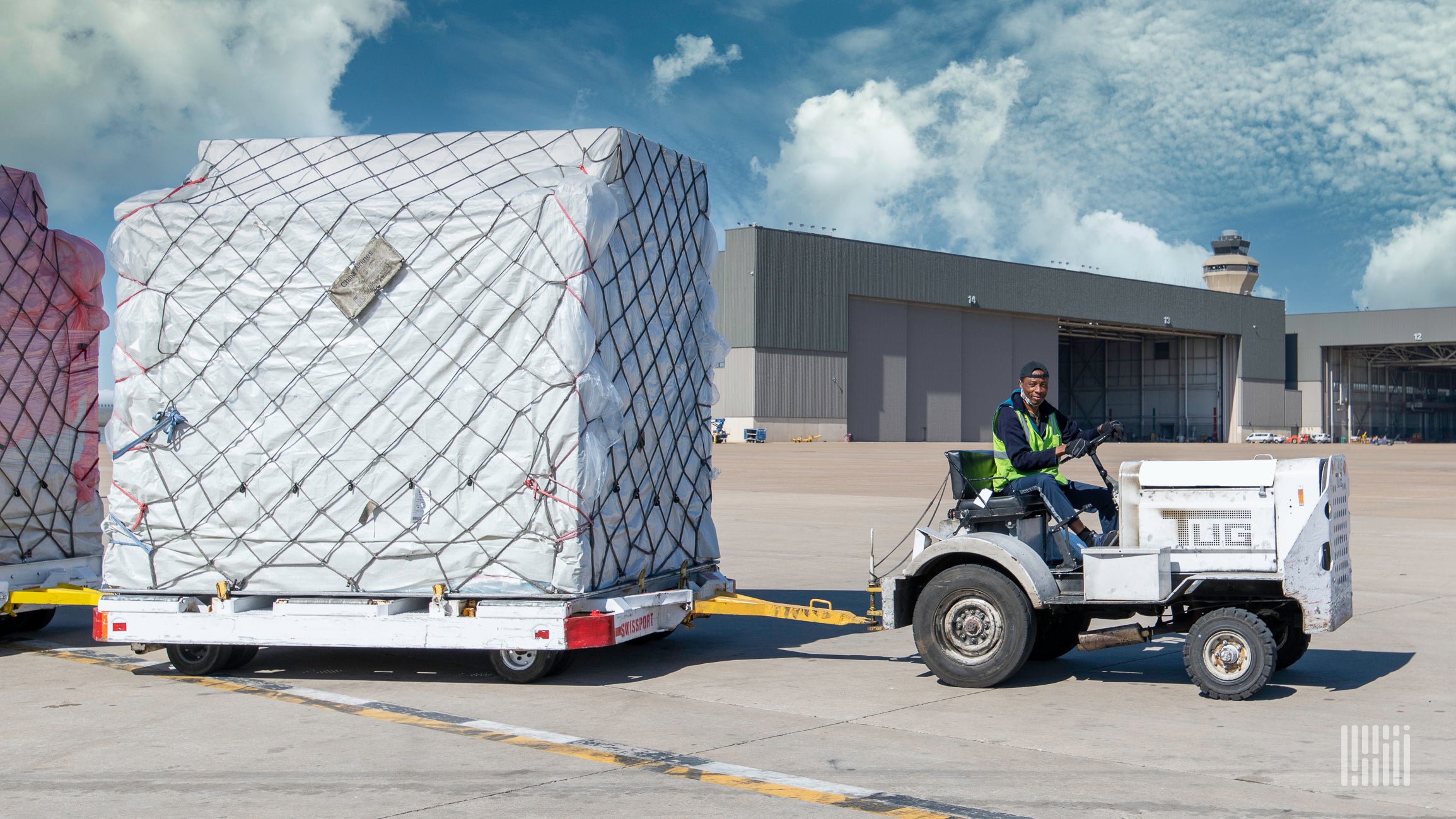 A large air freight container on a flat cart pulled by a tug.