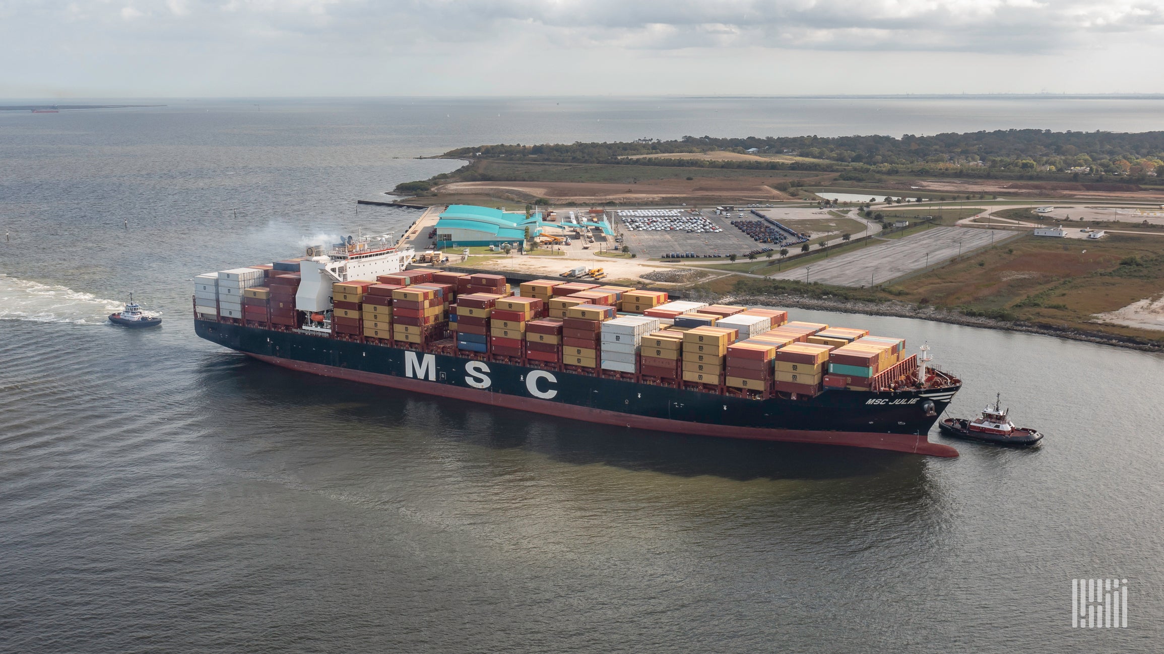 A massive container ship with MSC logo is pulled by tugs into the ship channel.