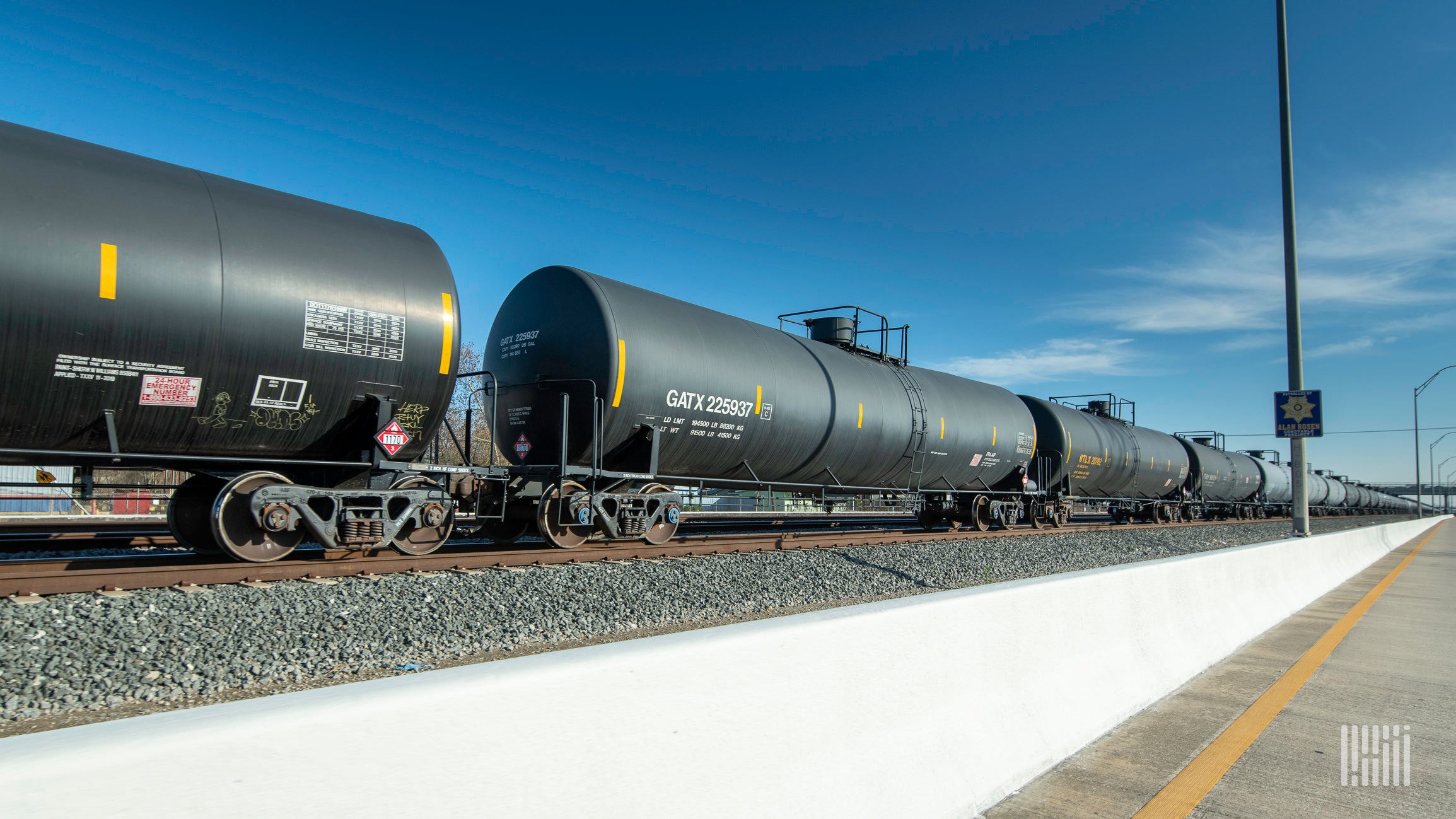 A line of tank cars moves on railroad track on a bright day.