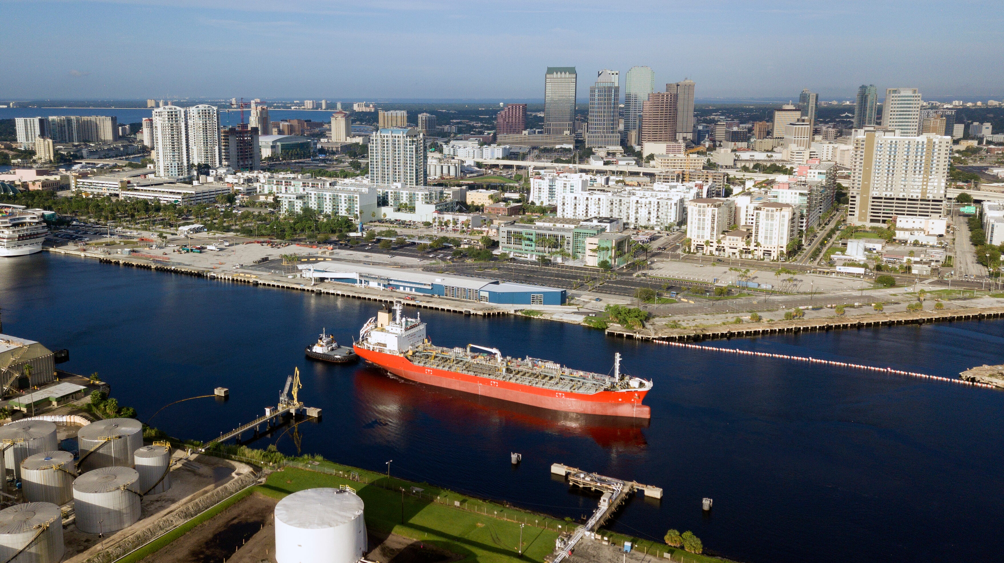 An oil tanker vessels sails into a harbor. A city landscape of tall buildings is in the background.