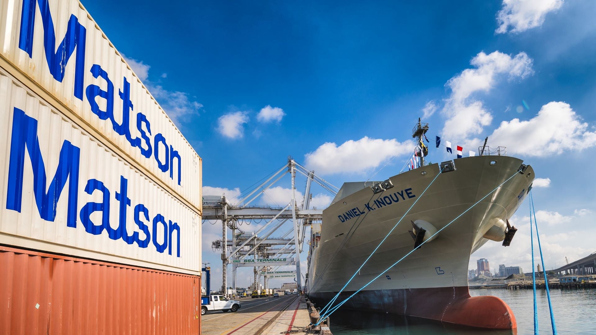 A view of a large cargo ship at the dock, looking straight on from the bow. with Matson containers stacked on the dock to the side on a bright day.