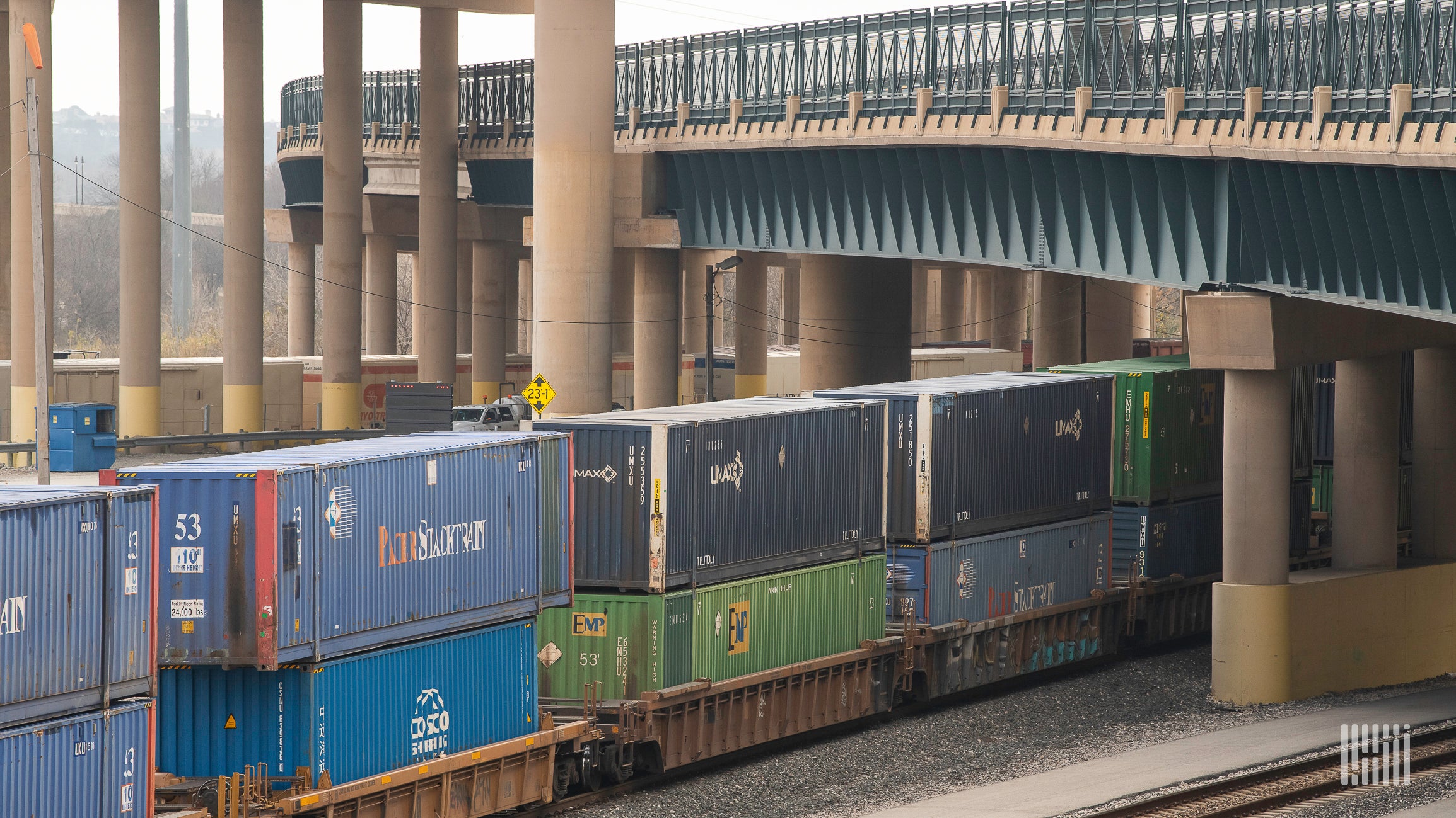 A train hauling intermodal containers passes by under a bridge.