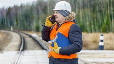 A man wearing a hard hat and gloves stands by a rail track while talking on a mobile phone.