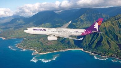 A white Hawaiian Airlines jet with blue tail flies over a lush green island and water.