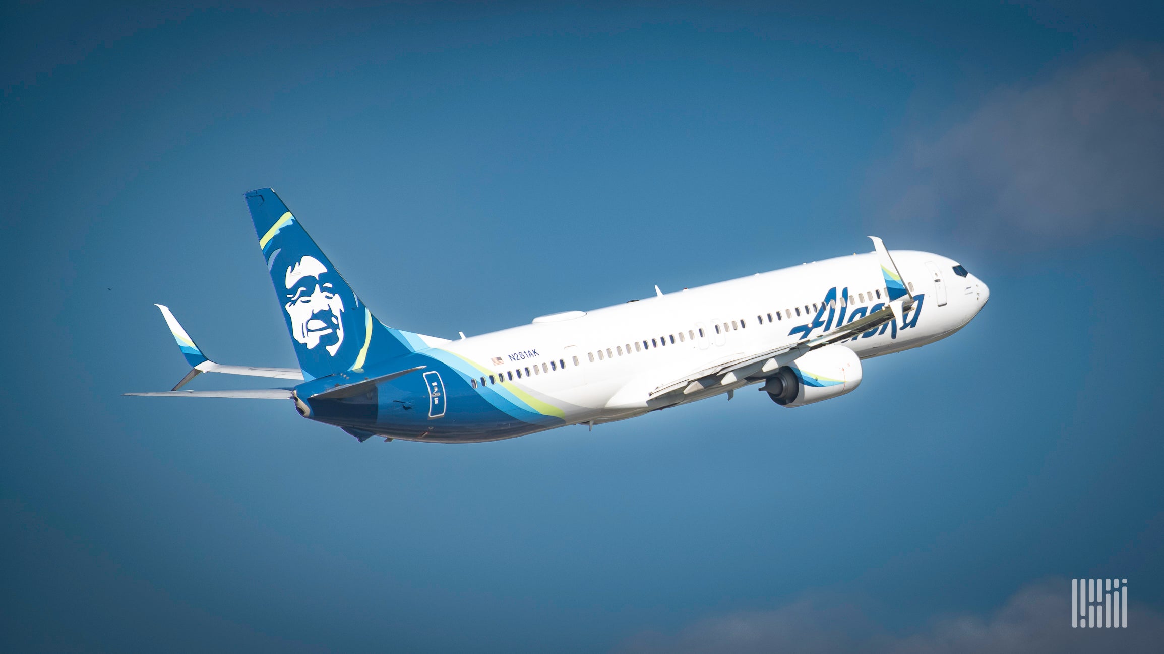 A white jet with aqua tail and Alaska Air logo flies high into a blue sky, as seen from the side rear.