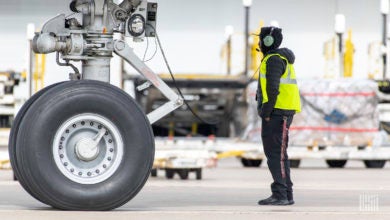 An airport ramp worker evaluates the wheel of a large cargo jet with a pallet of freight in the background.