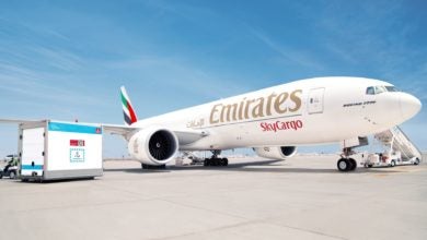 A white Emirates cargo jet with gold lettering and a container sit on the tarmac on a sunny day.