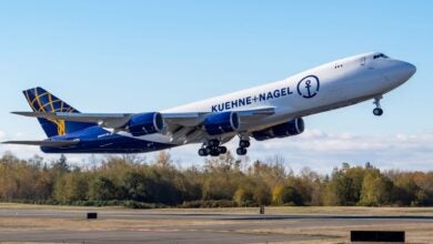 An Atlas Air jumbo jet with blue rear and white front half and Kuehne + Nagel logo takes off from a runway with trees in background.