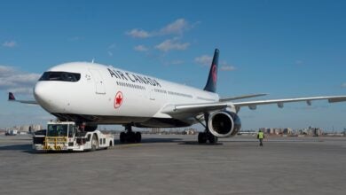 A white Air Canada jet with black tail being pulled by an airport tug on a sunny day.