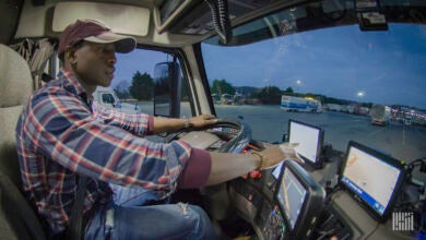 Man sits behind the steering wheel in the cab of a semi truck, touching the screen of a tablet on the dashboard