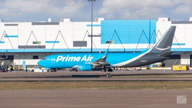 A light blue plane with Prime Air on the side rolls by a white and blue warehouse at the airport.