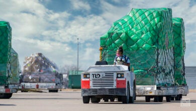 An airport cargo tug with a woman driver with black hair pulls a pallet with green shrink wrap on a bright day.