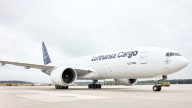 A white jet with blue tail and lettering "Lufthansa Cargo" on the tarmac, viewed from side ground level.