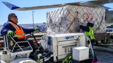 A man with an orange vest drives a cargo tug up to an aircraft to load a pallet.