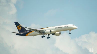 A brown-tailed UPS plane approaching airport with wheels down on a sunny day and clouds in the background.