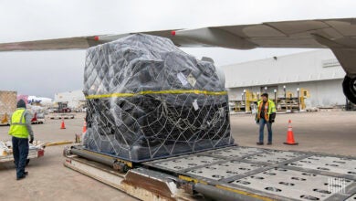 A large pallet, shrink-wrapped in black plastic rests on the tarmac under the wing of a plane.