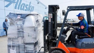 A forklift drive moves a pallet of items wrapped in plastic next to an Alaska Air warehouse.