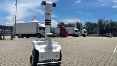 A two-wheeled robot with cameras in a warehouse parking lot on a sunny day, with white trucks in the background.