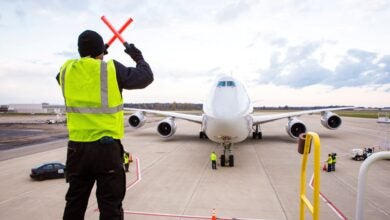 A man with a yellow vest and orange batons signals to pilots in a large jet on how to approach the gate.