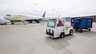 A blue-tailed Delta jet in the background with stairs up to the cabin door, and a tug pulling a blue baggage cart in the foreground.