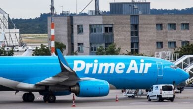 A light blue Amazon Prime Air plane parked at an airport with buildings in the background.