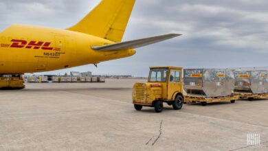 Tail end of a mustard-colored aircraft with a yard truck pulling carts with containers at an airport.