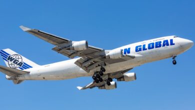 A white jumbo jet with blue lettering for Western Global, seen from below with wheels down on approach to airport with blue sky in background.