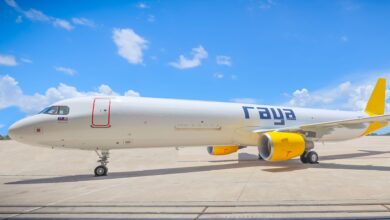 A yellow-tailed jet with yellow engines and "Raya" lettering sits on the tarmac under a bright blue sky in a close up view from the side.