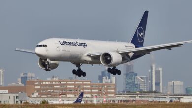 A blue-tailed Lufthansa Cargo jet approaches the runway with city buildings in the background.