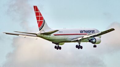 A red-tailed cargo jet flying with wheels down near an airport, viewed from behind.