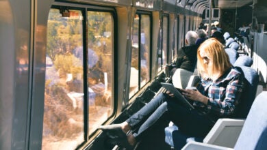 A woman sits next to a window of a train with a laptop and a cup of coffee in her hands.