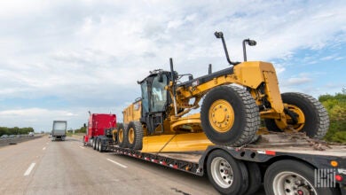 A red tractor pulling a motor grader on a flatbed trailer