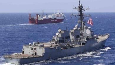 A U.S. Navy destroyer seen from the rear moving at sea with a red commercial vessel off the port side.