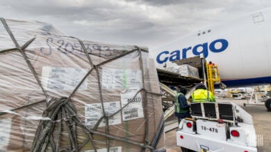 A large pallet of freight covered in plastic and straps on the tarmac next to a large cargo plane.