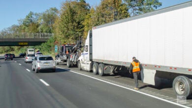 Semi-truck on the side of the highway with a driver in an orange vest walking next to it.