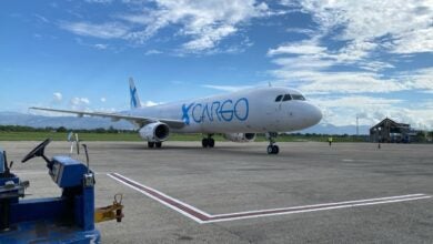 A GlobalX cargo jet in light blue lettering on tarmac of small tropical airport as seen from ground level in front, blue skies above.