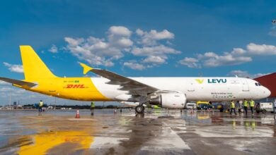 A half white/half mustard DHL freighter aircraft on a wet tarmac with a blue sky and white clouds in the background.