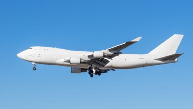 An all-white jumbo jet freighter on airport approach with wheels down, blue sky behind it.