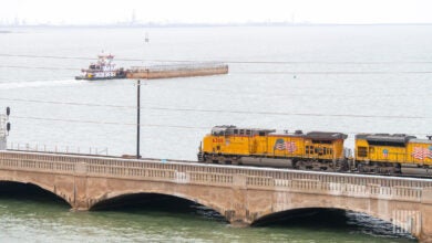 A yellow Union Pacific freight train going over a low bridge with a tugboat pushing a barge in the background.