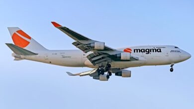 A Magma Aviation jumbo jet freighter with orange lettering seen from below as it comes in for landing with wheels down.