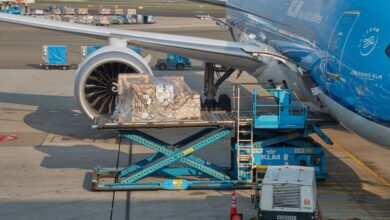 A light blue KLM jet is loaded with a large pallet of freight through a side door.