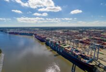 Image shows water, container ships, containers, cranes, blue sky.