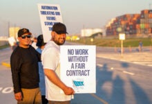 Image shows striking dockworkers holding signs with containers in background.